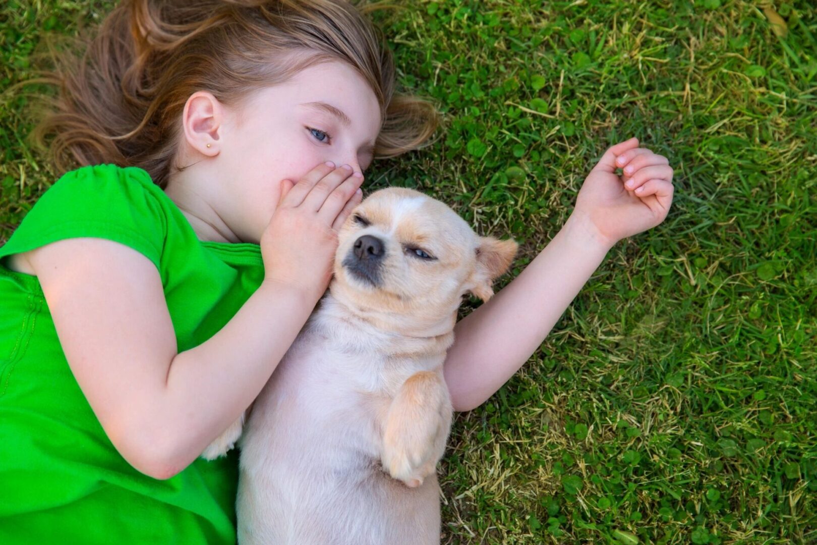 A little girl laying in the grass with her dog.